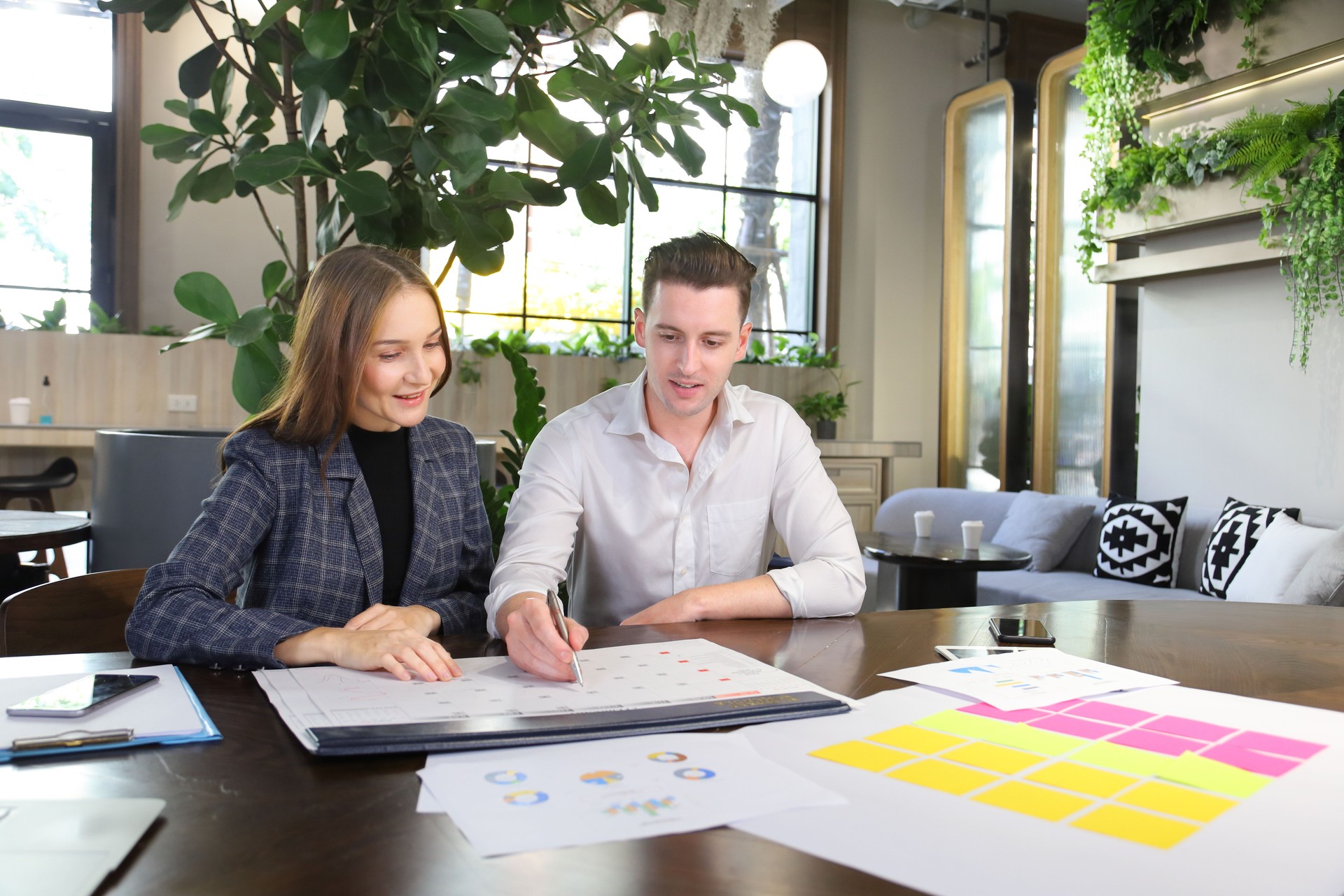 Kaukasische Geschäftsleute arbeiten an einer neuen strategischen Planung für das nächste Jahr mit Kalender, während sie in einem umweltfreundlichen, modernen Arbeitsraum arbeiten, der von einer luftreinigenden Anlage umgeben ist.